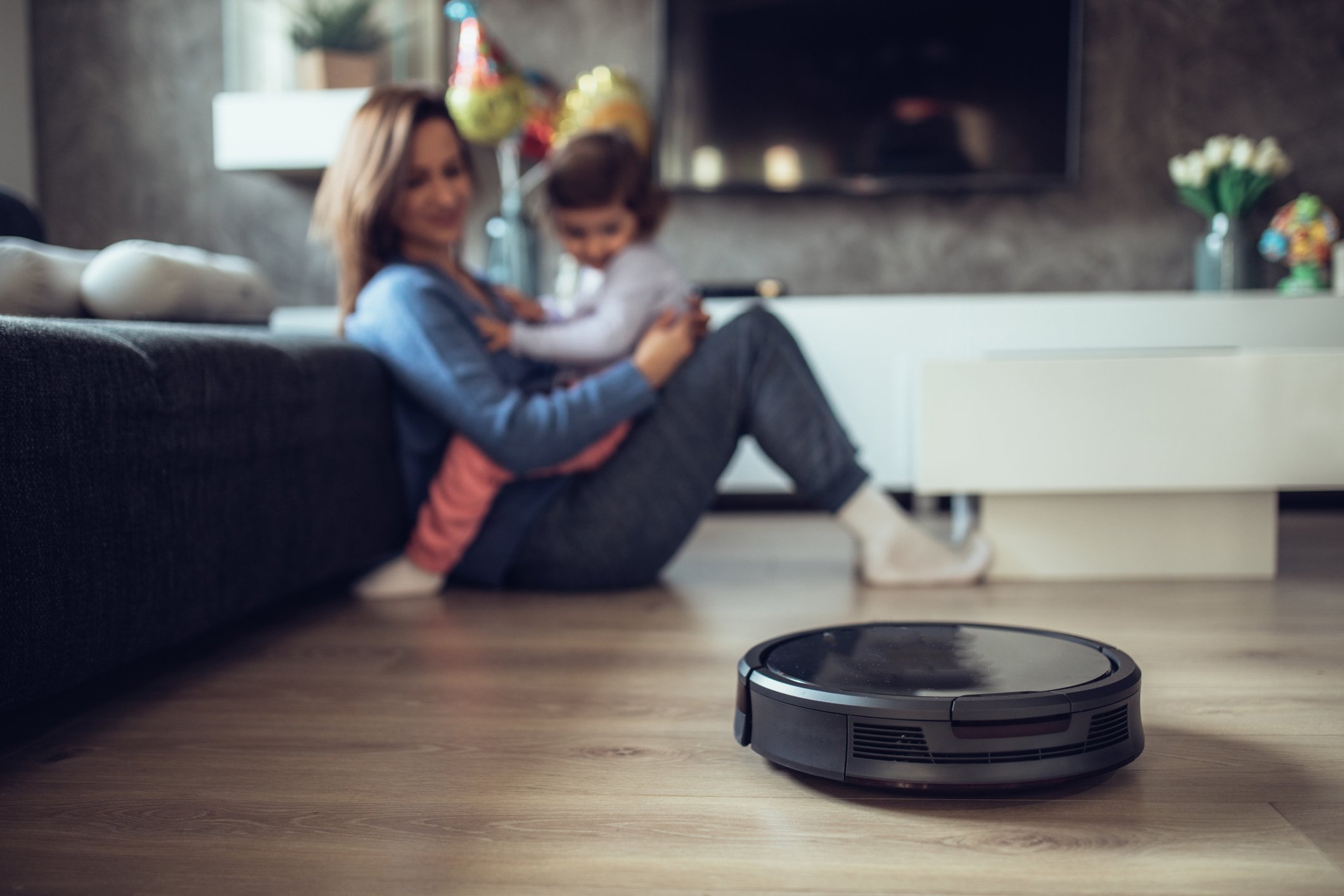 A mother and baby play on the floor while a robotic vacuum does its work.