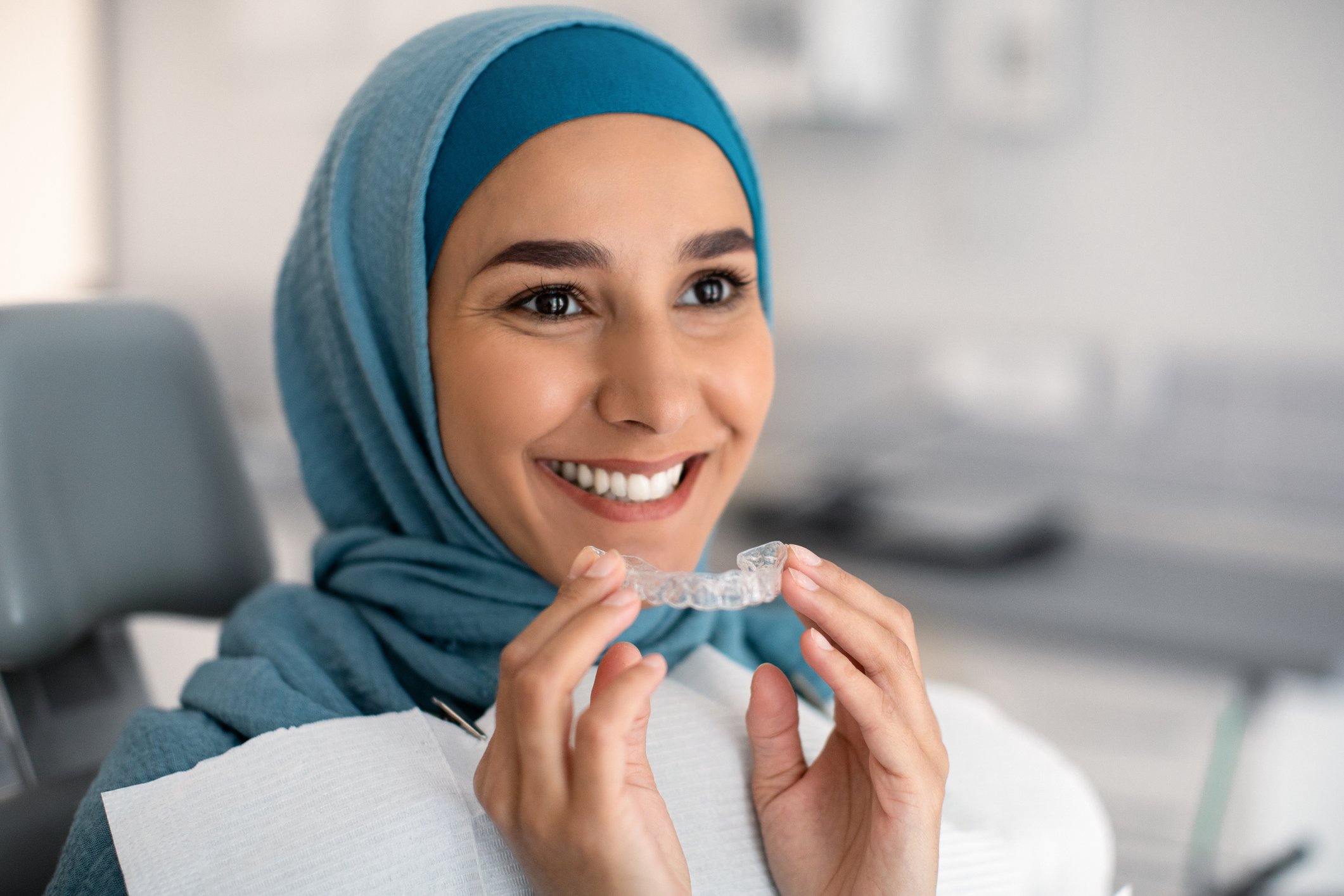 A person in the dentist chair holding a tray of clear teeth aligners while smiling.