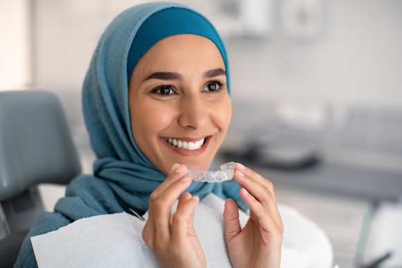 A person in the dentist chair holding a tray of clear teeth aligners while smiling.