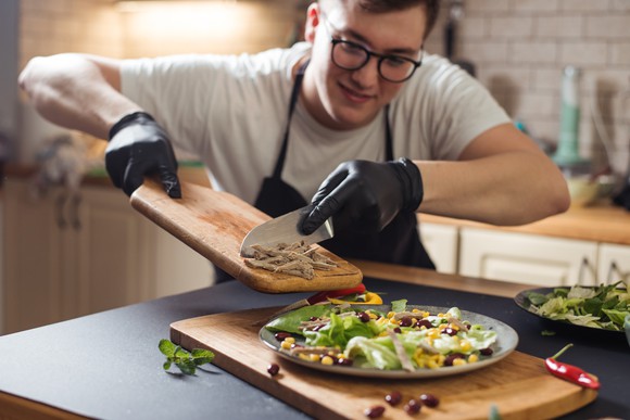 A person cooking a meal and sliding plant-based meat onto a dish of vegetables from a cutting board.