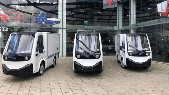 Three Cenntro Metro ECV vans parked in front of one of the company's buildings, with various corporate logos visible in the windows.