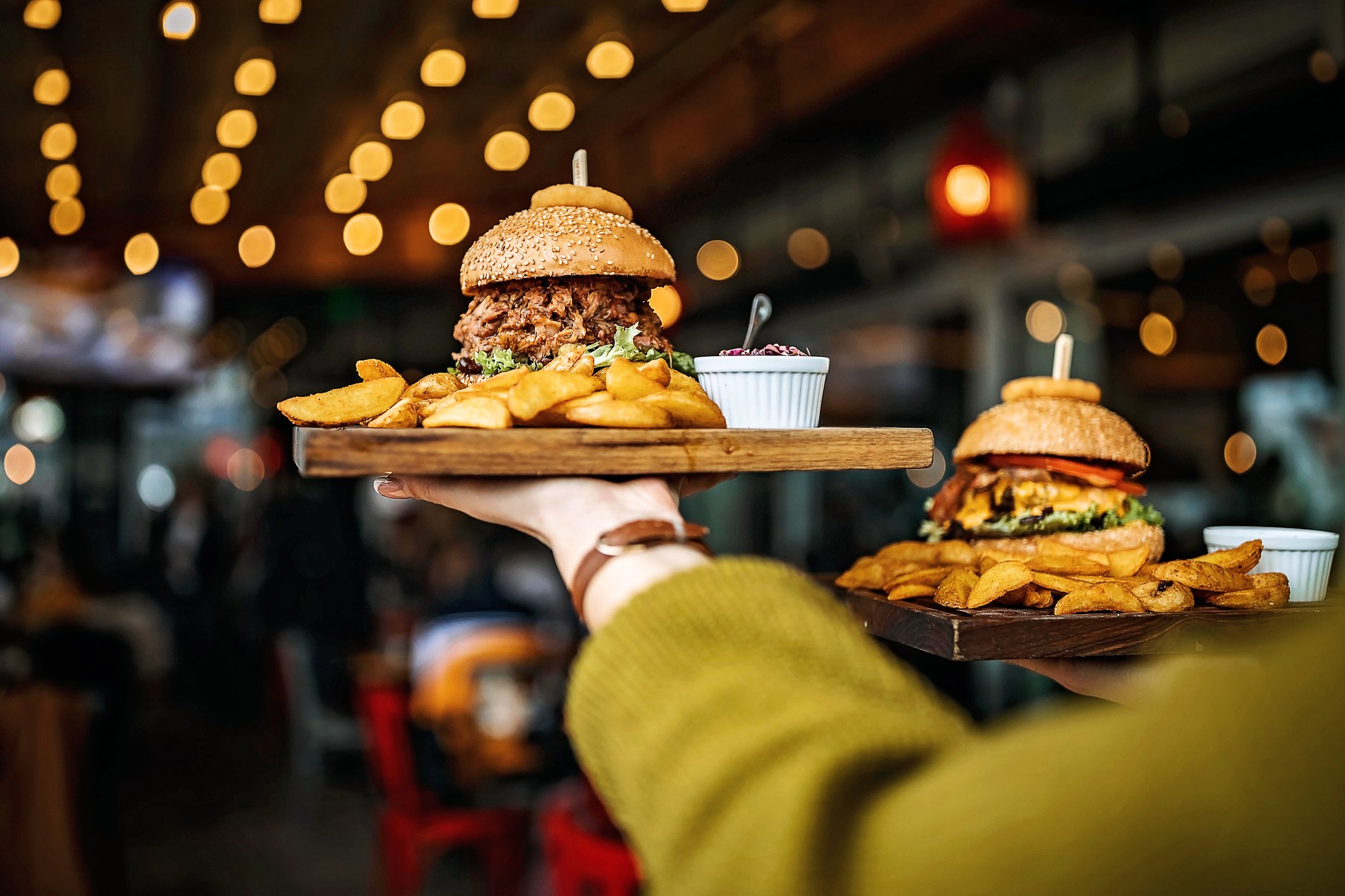 Two burgers loaded with toppings being brought to a table in a restaurant.