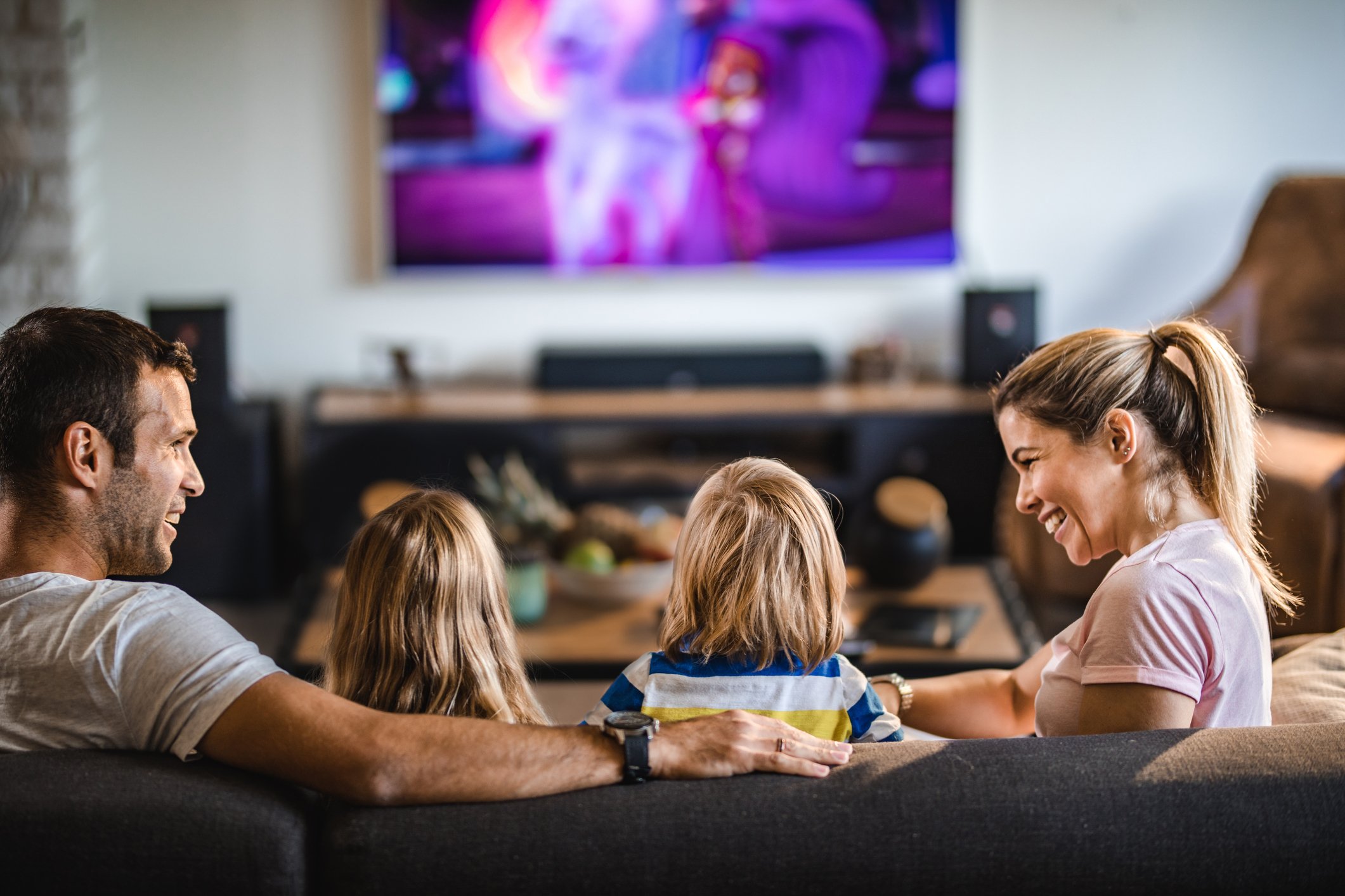 A family of four sits on a couch interacting while watching a streaming TV show on the television in the background