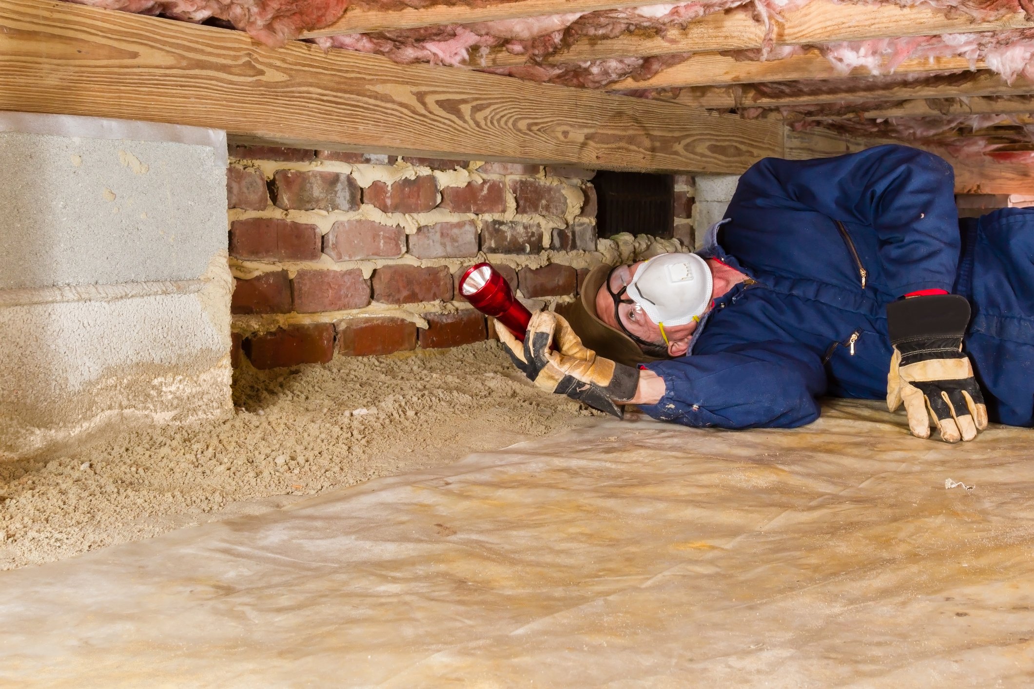 A person inspects a crawl space with a flashlight, wearing protective gear.