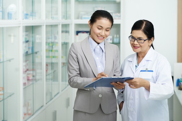 A pair of pharmacists consult a tablet while standing in the back of a pharmacy.