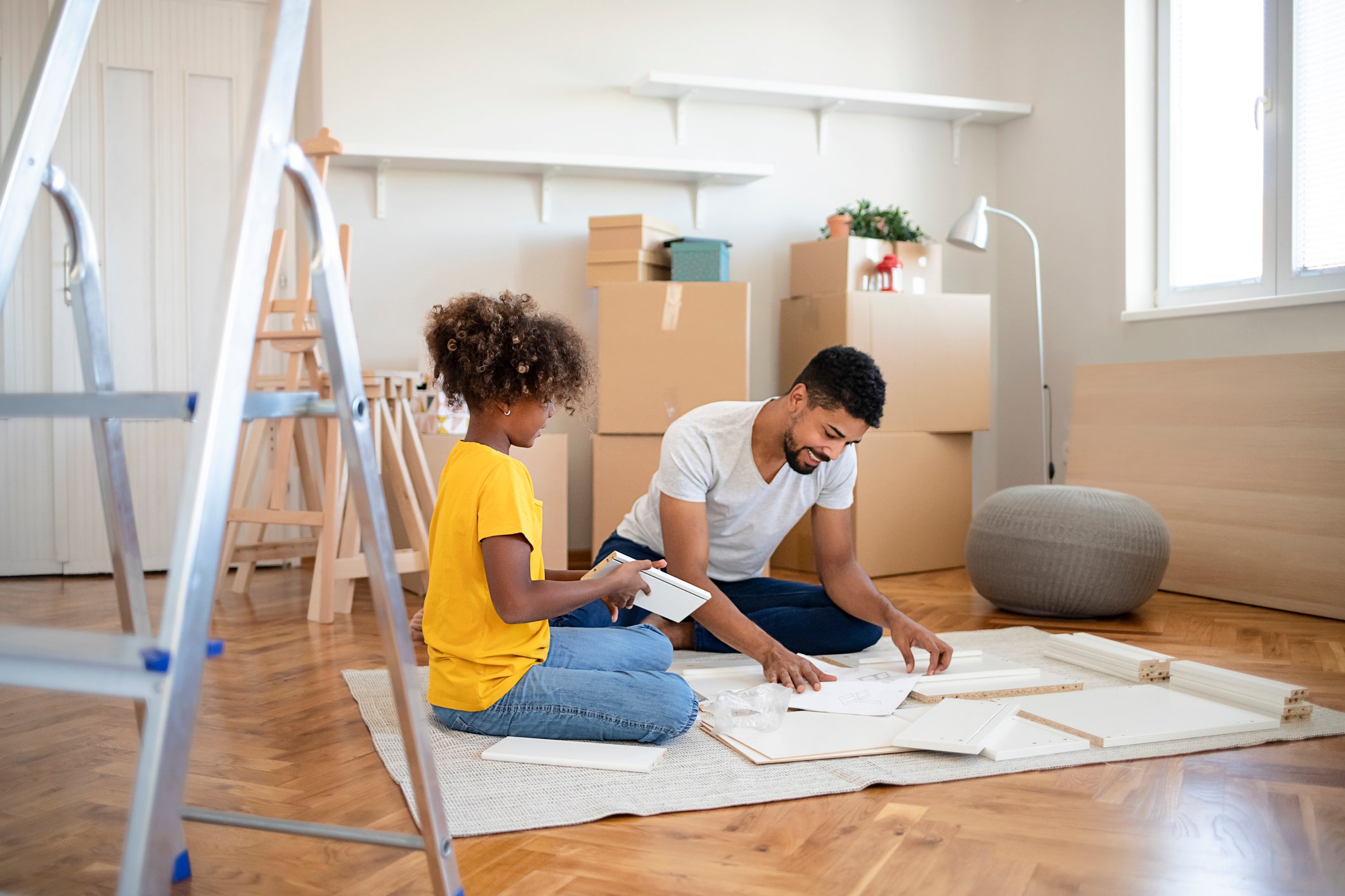 An adult and a child sit on the floor to assemble furniture in a new home.
