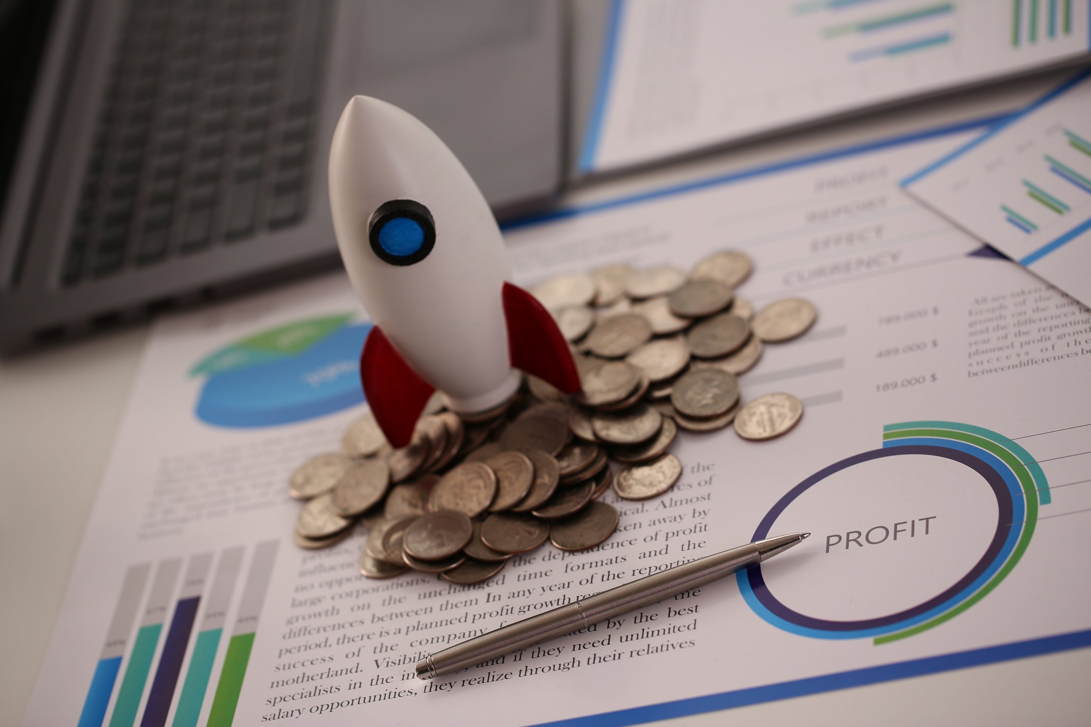 A toy rocket standing on a pile of coins in front of a laptop computer.