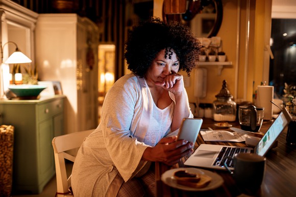 A person at their kitchen table, looking at their phone and open laptop, deep in thought. 