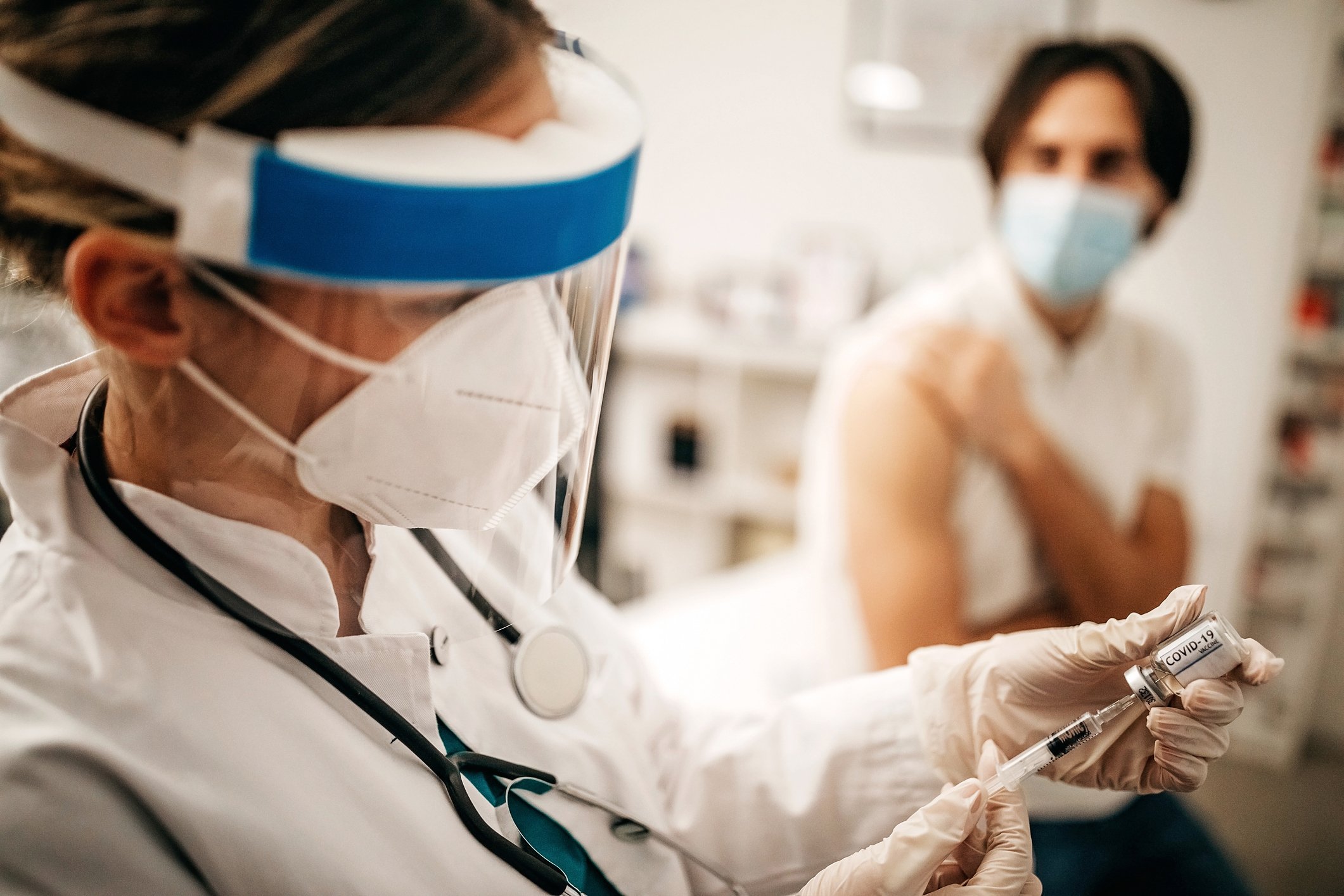 A healthcare worker wearing protective equipment loads a syringe with a dose of coronavirus vaccine while a patient waits in the background.
