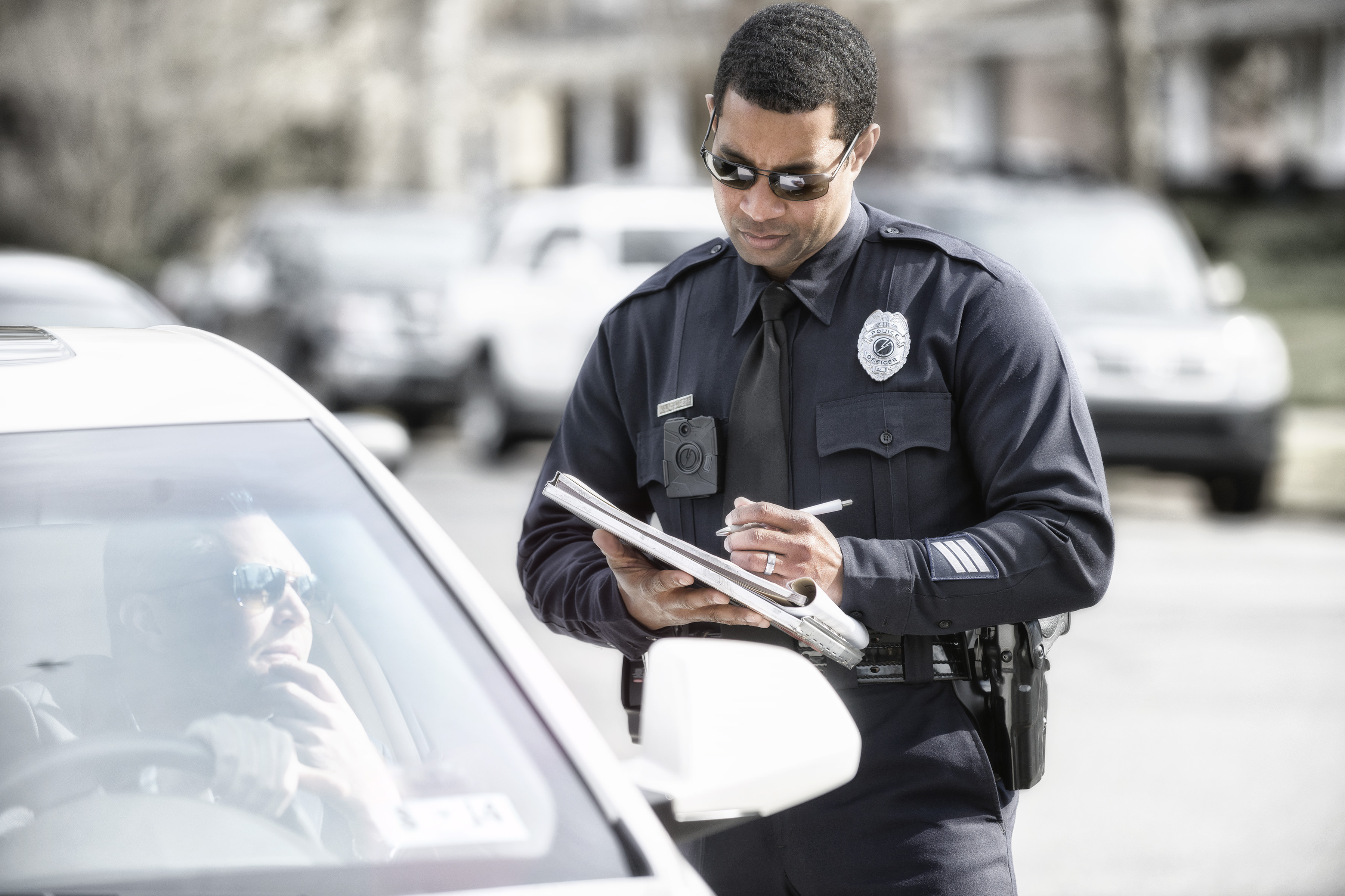 A police officer wearing an Axon Enterprises body camera holds a clipboard and pen while standing next to a vehicle as the driver of the vehicle speaks to the officer through the window