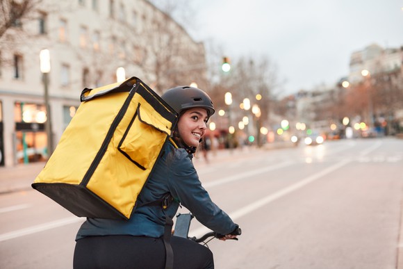 A food delivery courier in a European city at dusk, on a bicycle with a food bag and lights sparkling in the background. 