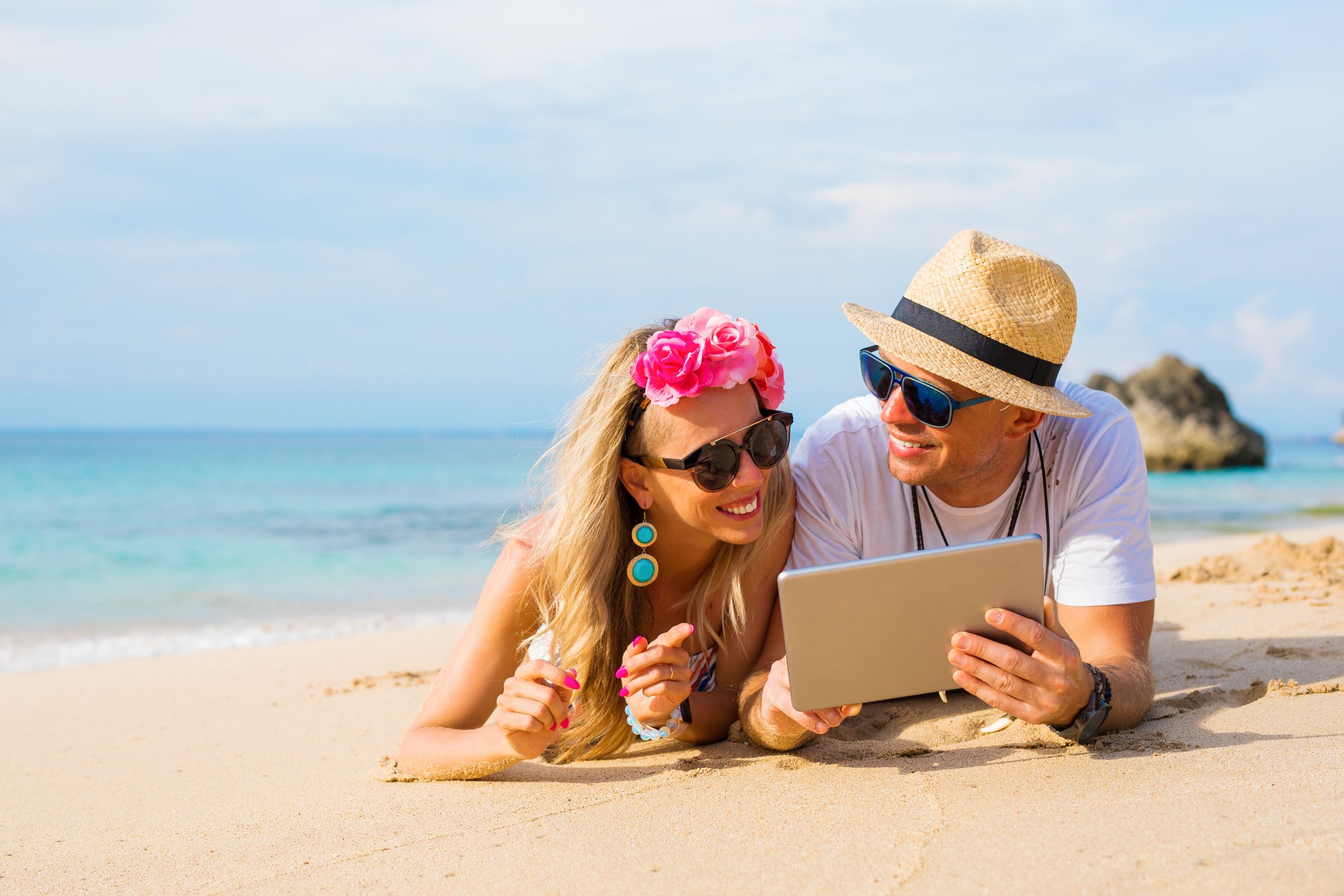 Two smiling people lying on a beach and looking at a touchscreen tablet.