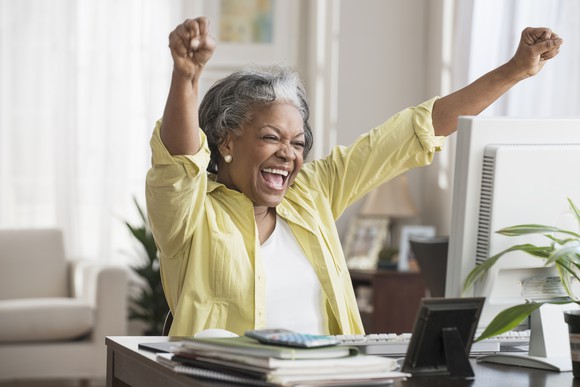 Senior celebrates with hands in the air at home, sitting at desk.