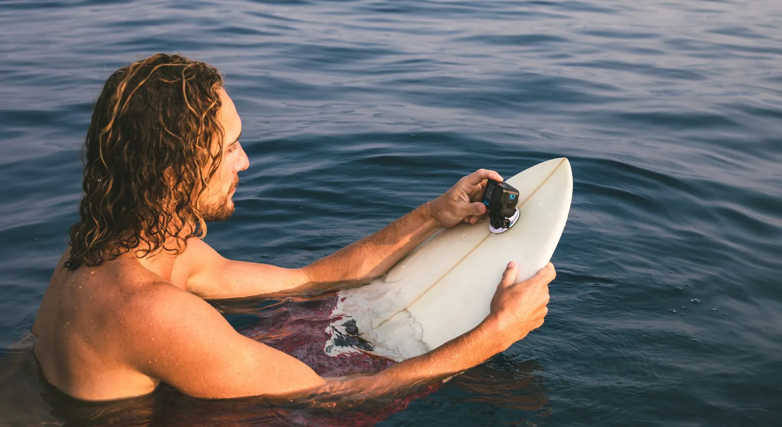 A surfer floats on a surf board in the water and makes adjustments to a GoPro camera attached to the surfboard.