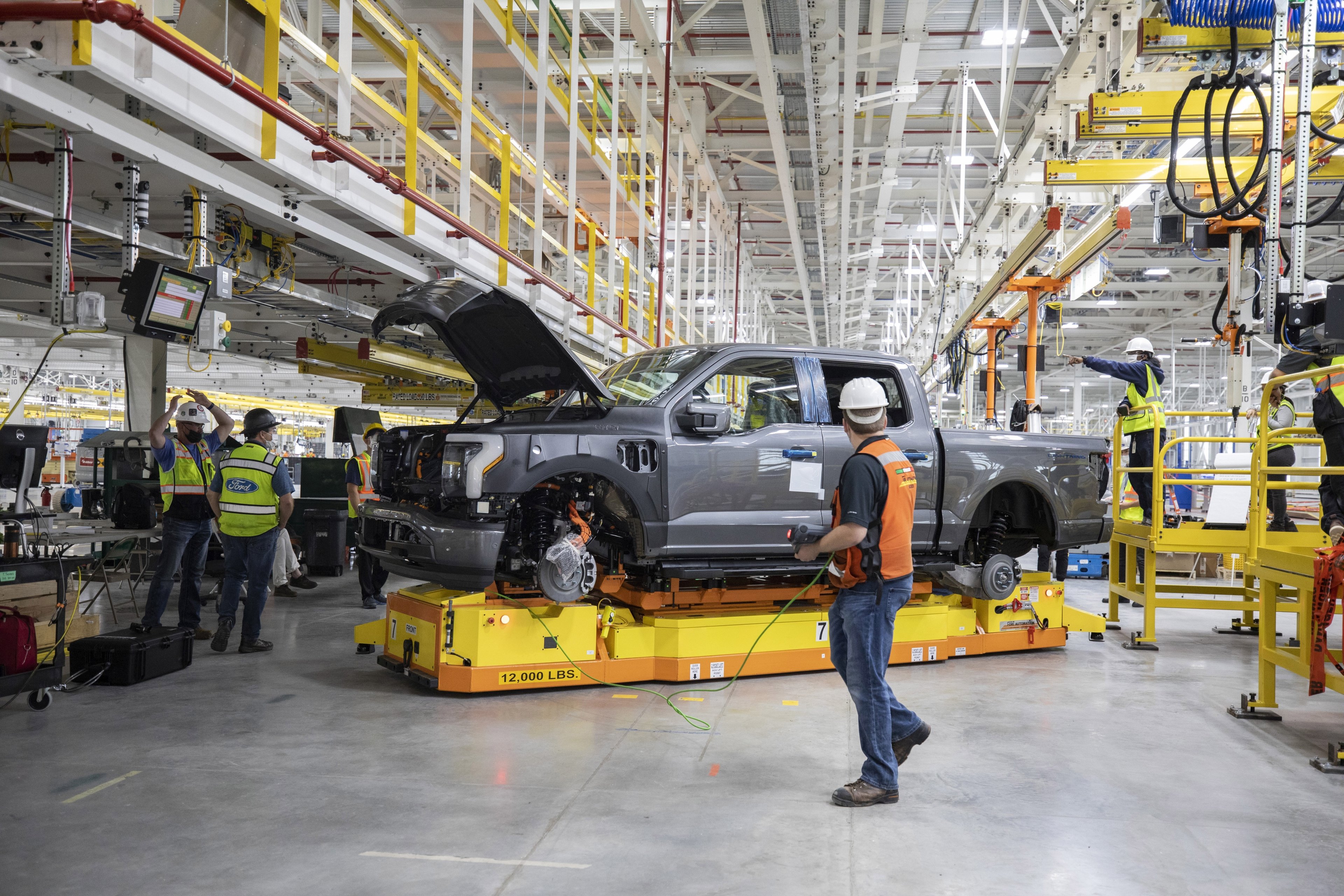 A pre-production Ford F-150 Lightning truck coming off the production line, surrounded by workers in hard hats. 