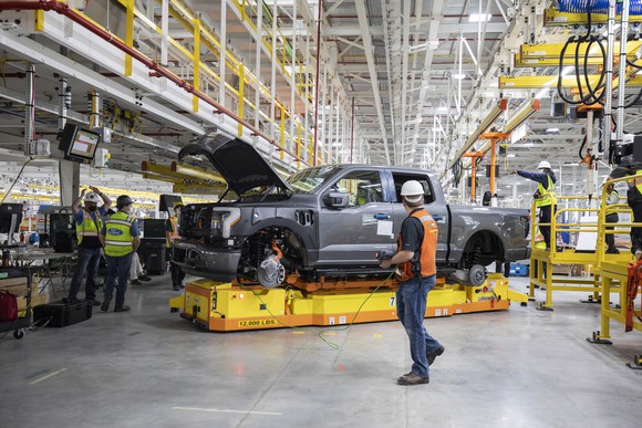 A pre-production Ford F-150 Lightning truck coming off the production line, surrounded by workers in hard hats. 