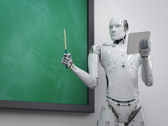 An artificial intelligence android stands in front of a chalkboard holding up a pointer and a tablet computer as though leading a class.