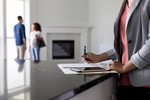 Two people stand in an apartment while a third does paperwork.