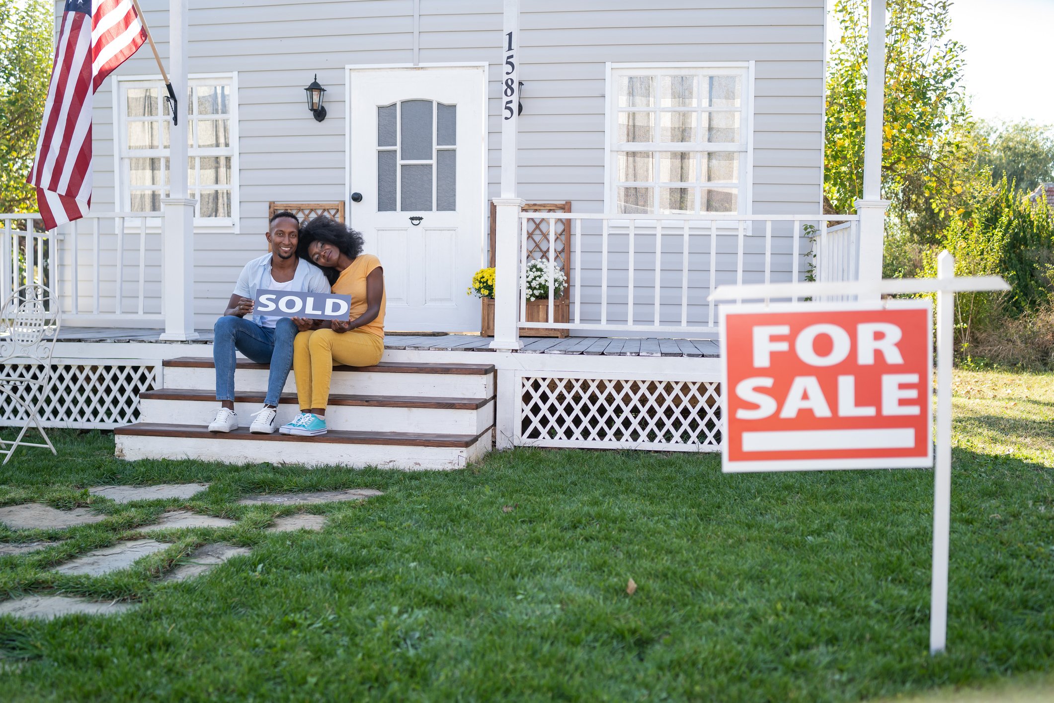 Two people sit on the porch of a home with a "For Sale" sign.  They hold a sign that says "Sold."