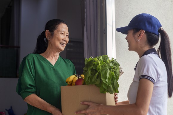 Grocery delivery in China with a young woman courier bringing a cardboard box of fresh groceries to an older woman at home. 
