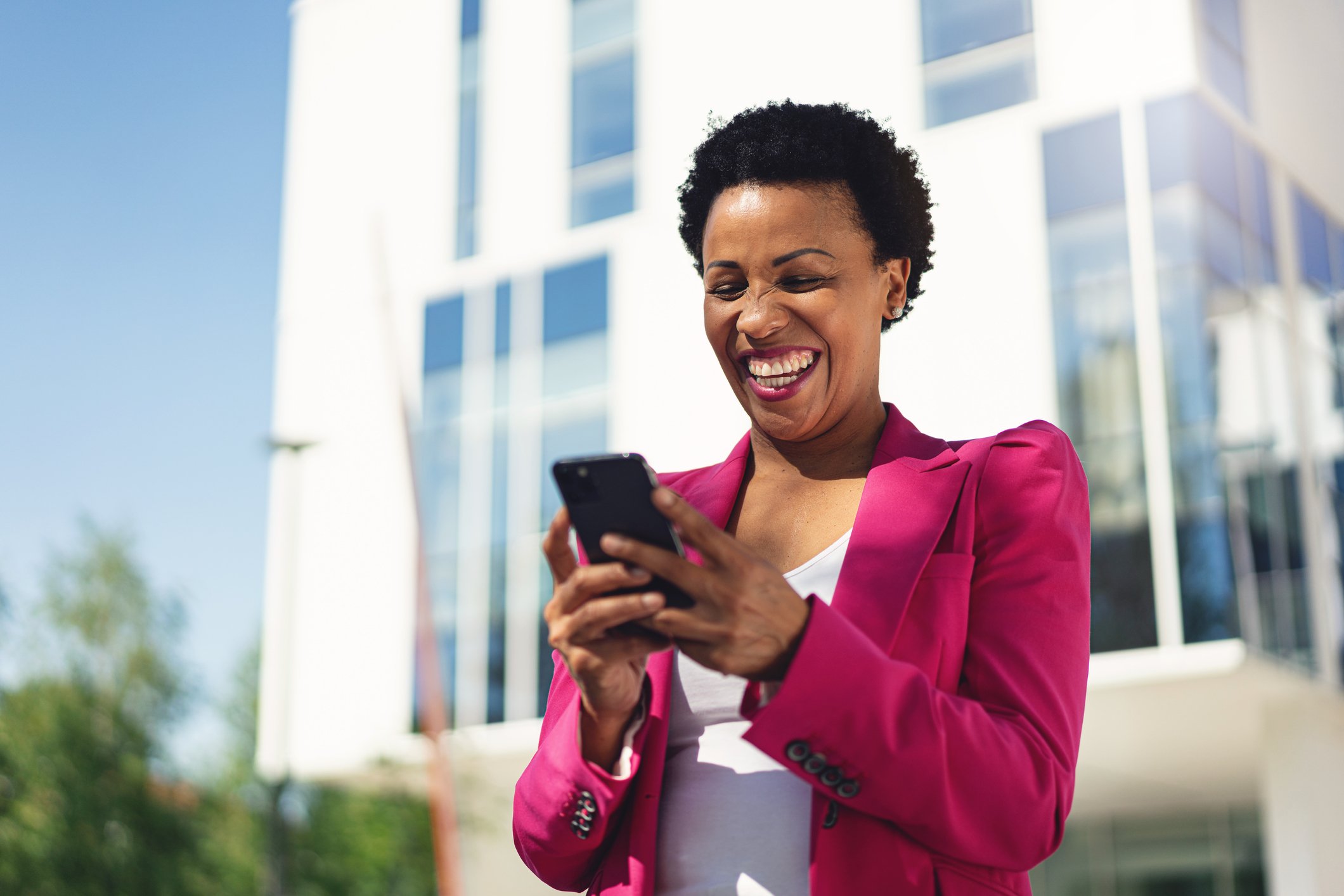 A businessperson smiles while looking at a smartphone in front of a building.