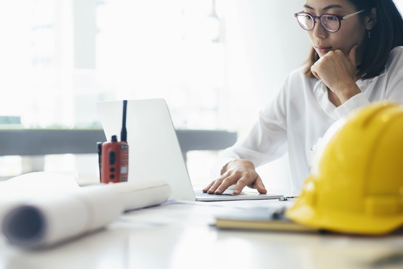 A person works at a laptop with a hard hat and blueprint in the foreground.