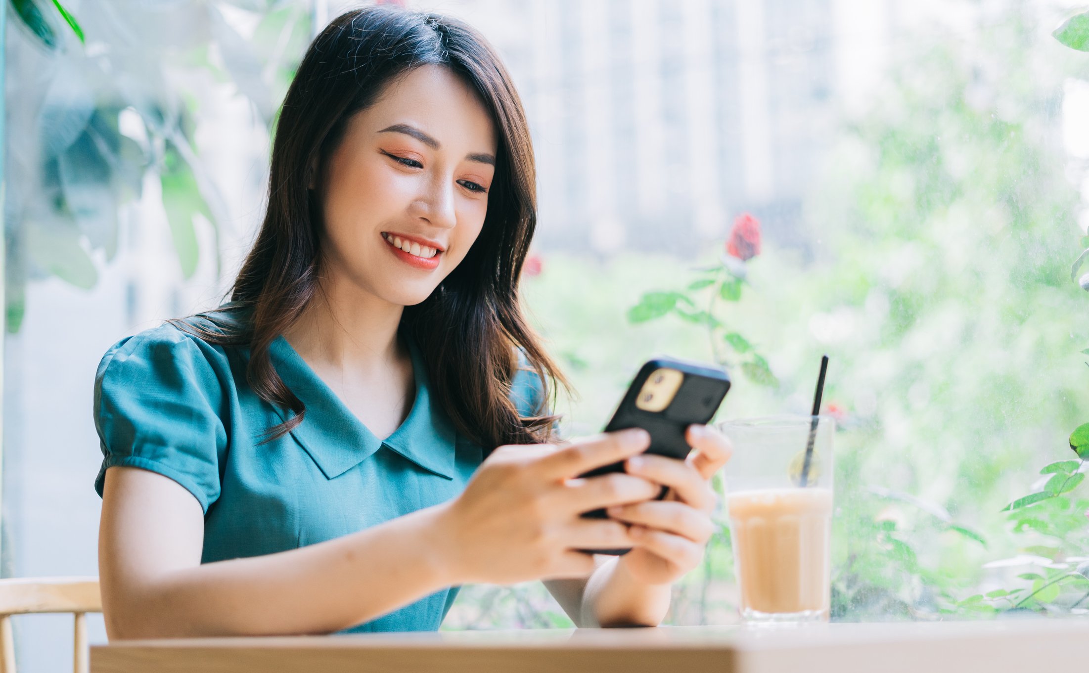 An Asian woman sits near a window at a table with a coffee sitting on it and looks at a smartphone she holds in her hand