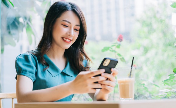 An Asian woman sits near a window at a table with a coffee sitting on it and looks at a smartphone she holds in her hand