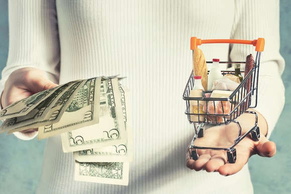 A woman weighing U.S. currency in one hand and a tiny shopping cart in the other.