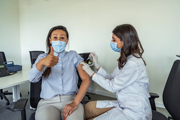 A patient gives an enthusiastic thumbs up while sitting in a chair and a nurse vaccinates her against the coronavirus.