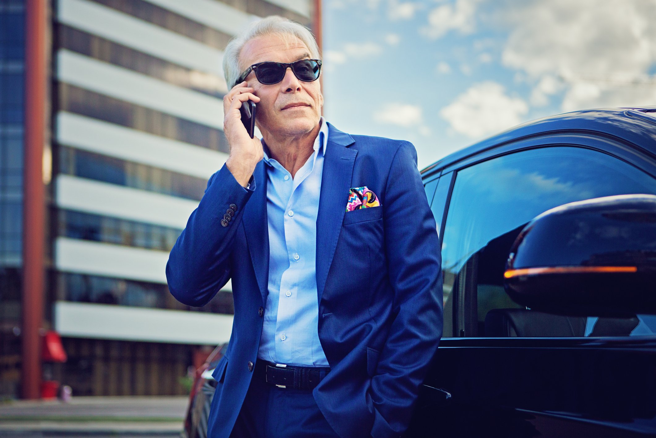 A business person holds a smartphone while standing in front of a car.