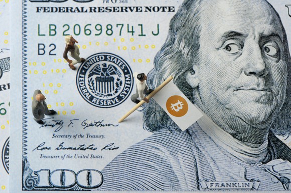 A small flag with the Bitcoin logo stuck into a hundred-dollar bill.