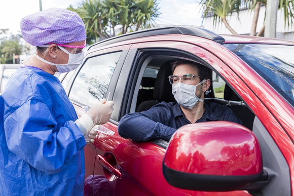 A man prepares to get tested for coronavirus while sitting in his car at a drive-through testing location as a nurse prepares a swab.