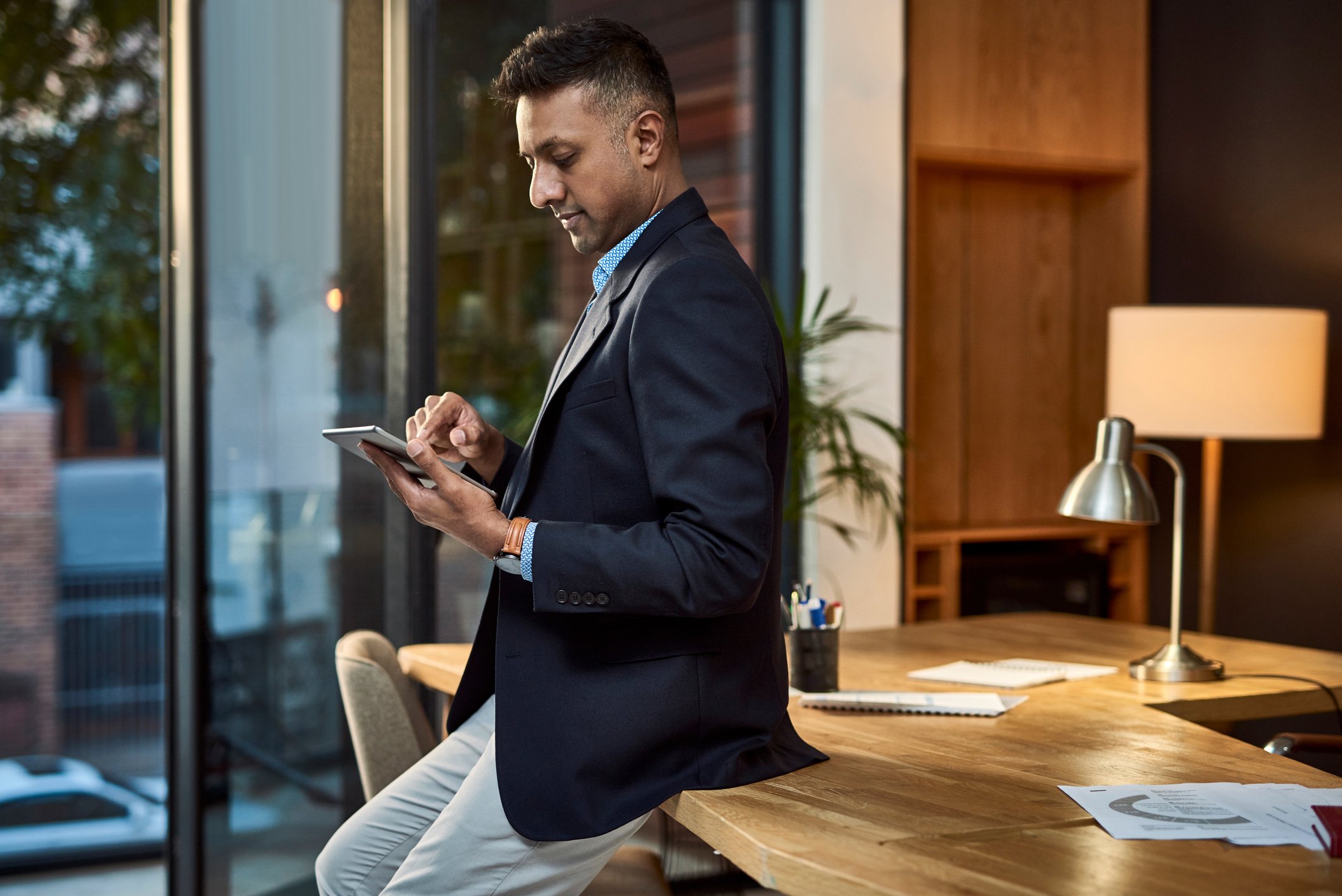Well-dressed individual sitting on edge of desk and looking at mobile device.