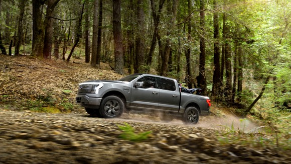 A gunmetal gray Ford F-150 Lightning Lariat electric pickup driving along an off-road trail through a forest.