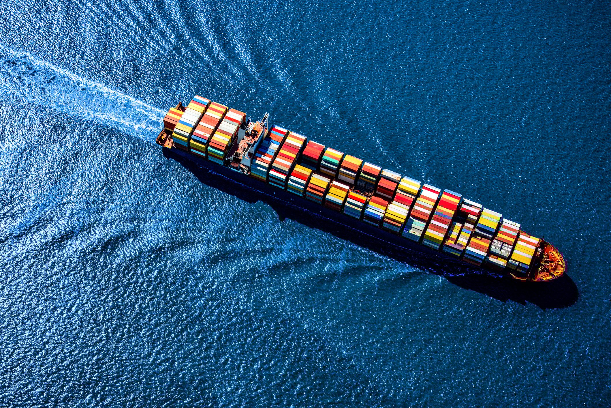 A huge container ship heavily laden with multicolored cargo containers, sailing across the blue surface of a sunlit ocean.