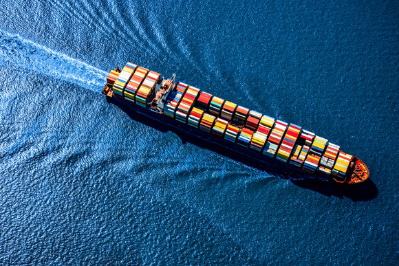 A huge container ship heavily laden with multicolored cargo containers, sailing across the blue surface of a sunlit ocean.