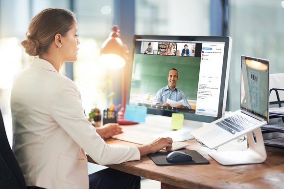 A businessperson videoconferencing with several people, using a laptop and desktop monitor.