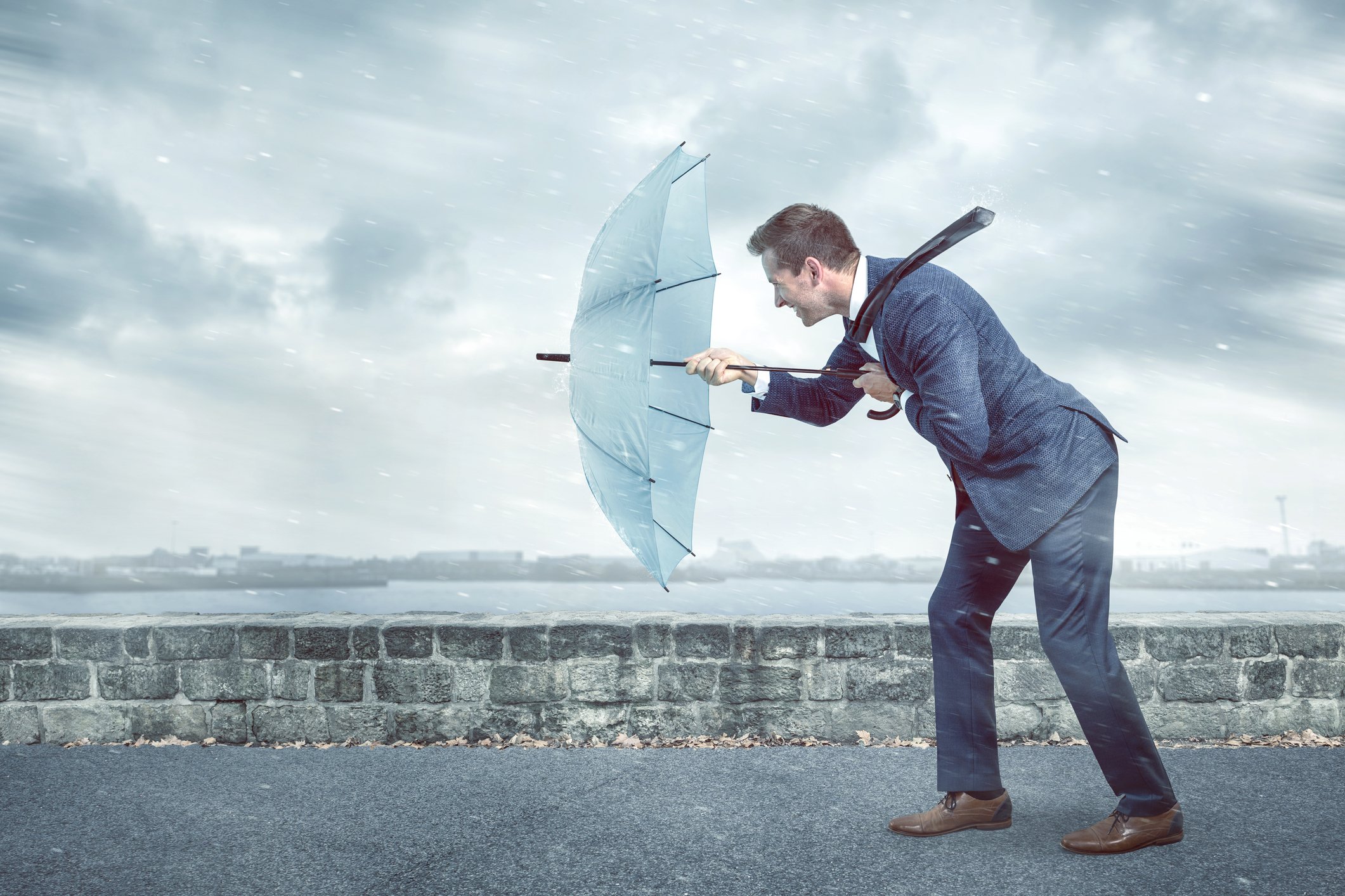 A business man holds out a light blue umbrella to brace against a strong headwind. 