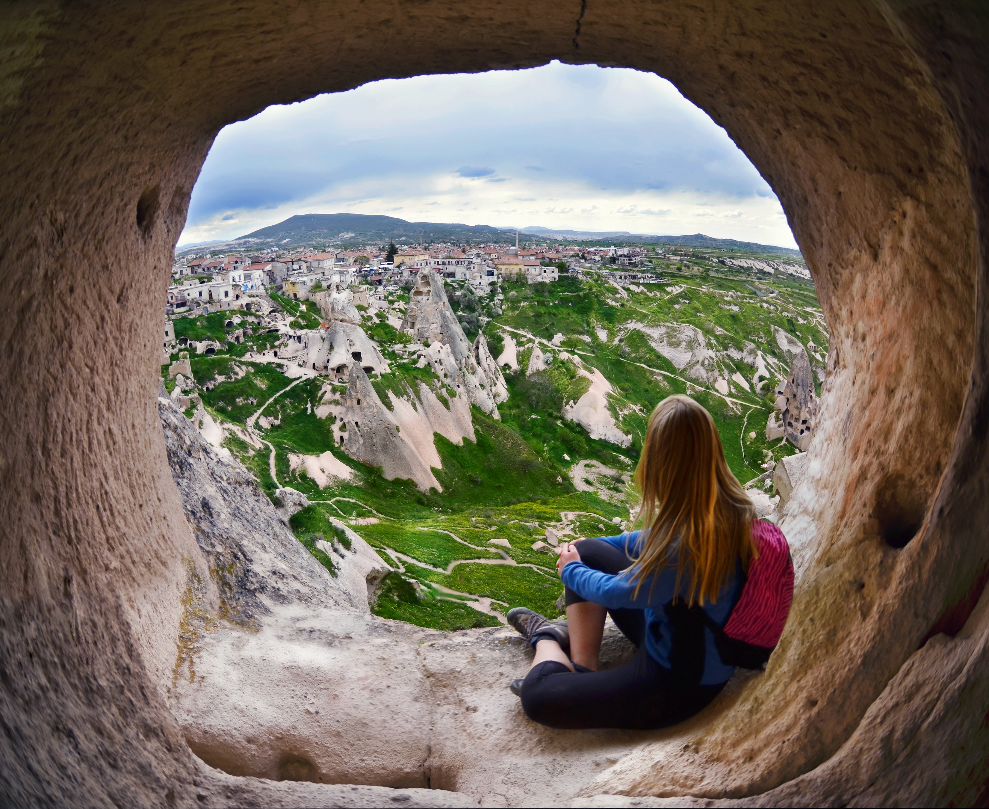 Person sitting inside a huge rock opening, looking out at a green and rocky landscape.