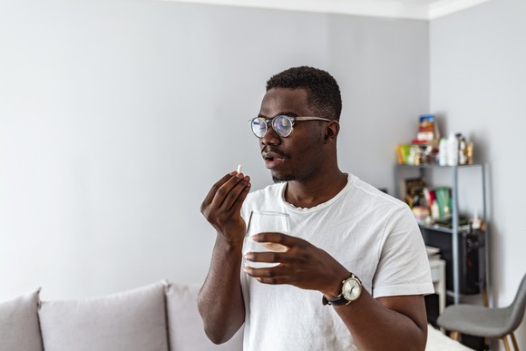 Patient taking a pill in a room with gray walls, a gray couch, and a shelf in the background. 