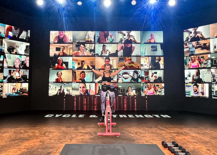 Trainer leading a stationary bike exercise session in front of many TV screens.