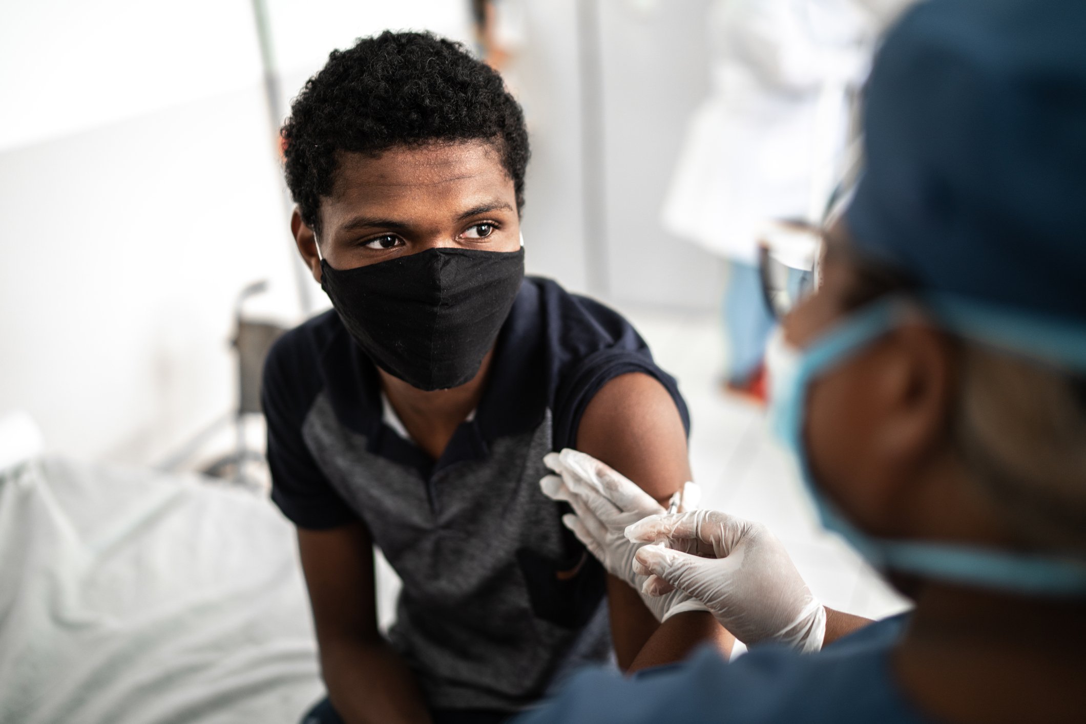 A doctor wearing protective equipment vaccinates a teenager sitting on a bed.
