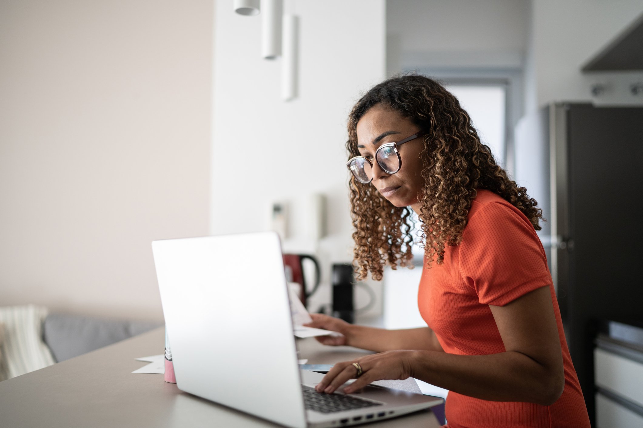 A person sitting at the kitchen counter looking at a laptop and bills.