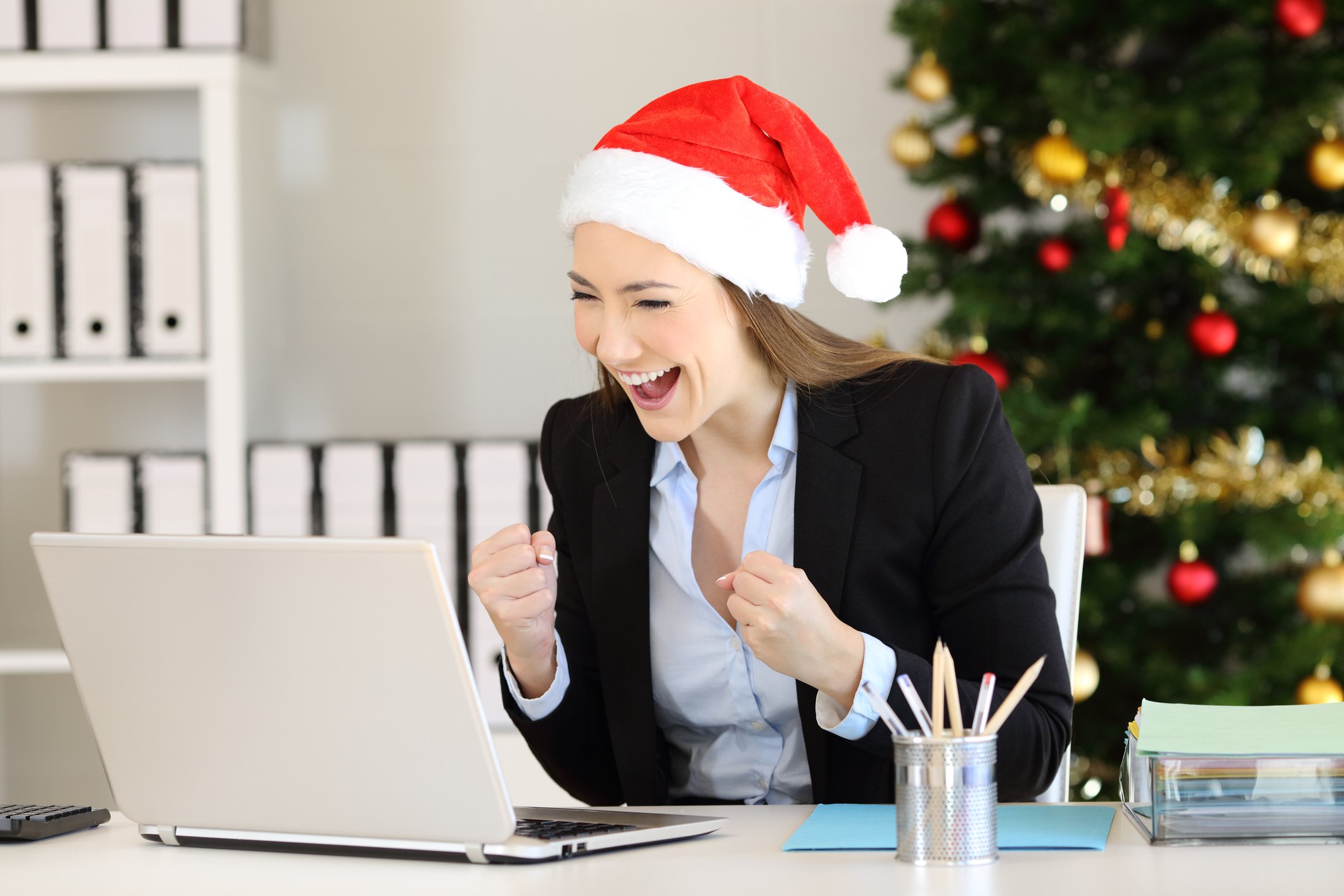 A smiling person wearing a Santa hat looking at a laptop.