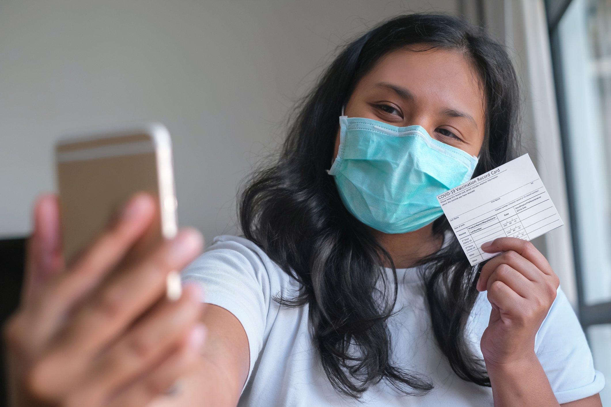 A masked woman takes a selfie with her vaccine card.