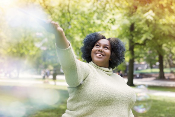 A woman smiling in a park with her arm raised as soap bubbles float around her. 