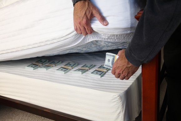 A person stashing stacks of cash under a mattress.