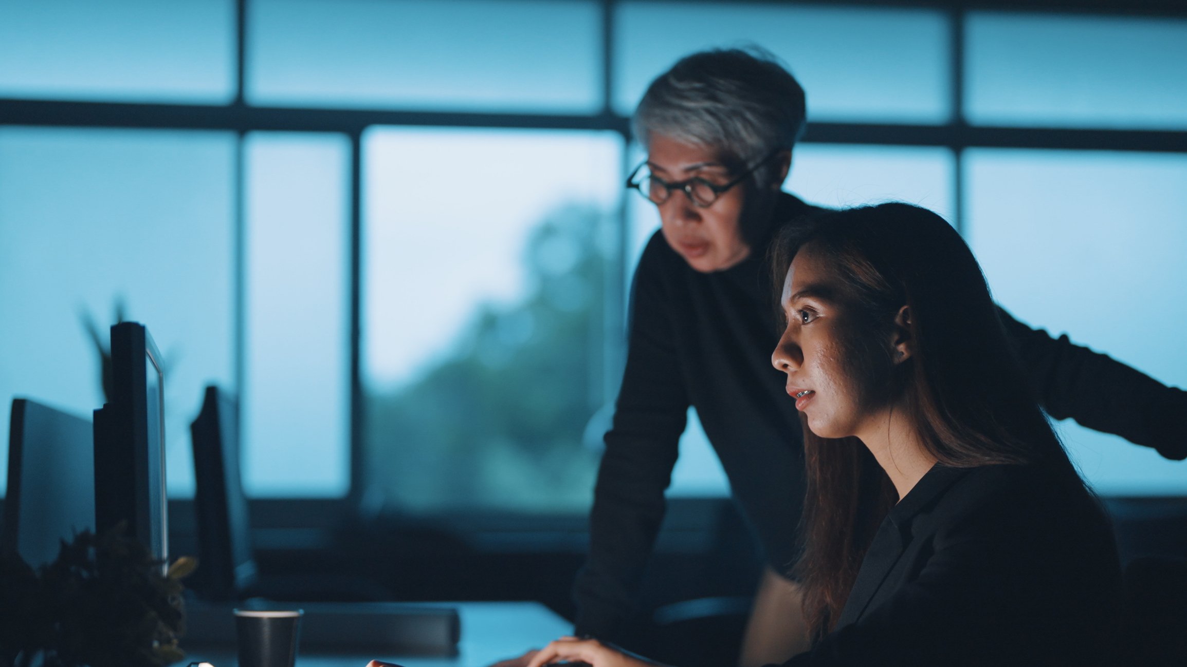 Two office workers working together at a desk with computers and monitors.