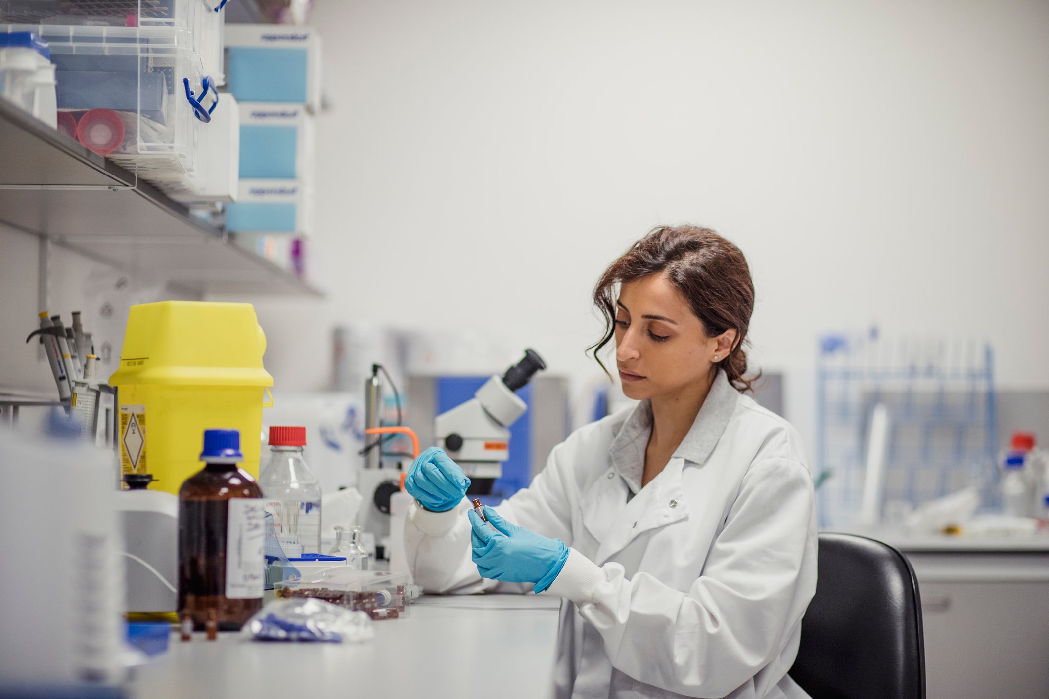 A scientist examines a vial while sitting at a laboratory bench.